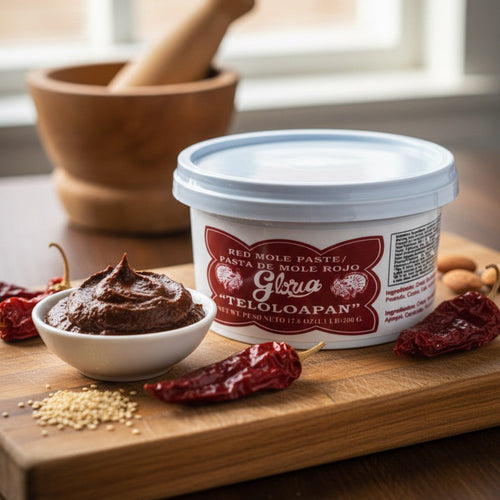 Container of Gloria Teloloapan mole paste on a wooden cutting board with dried chilies and a mortar and pestle.
